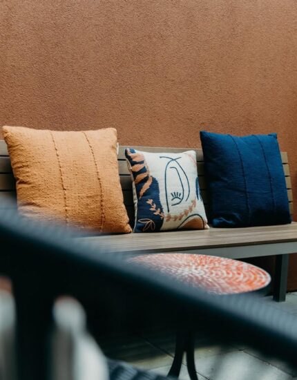 Three decorative pillows—orange, patterned, and navy—are arranged on a wooden bench against a brown wall, with part of a table and chair in the foreground.