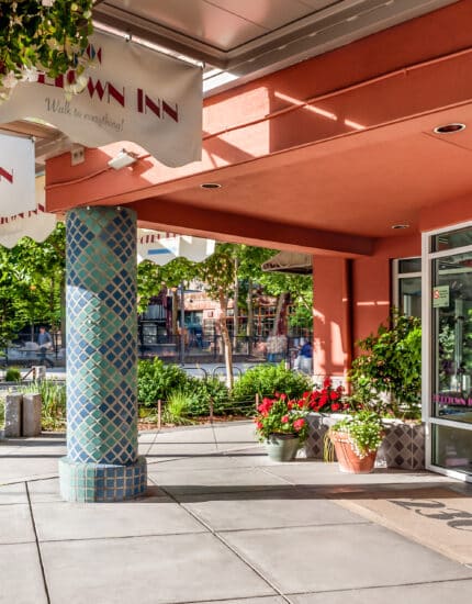 The entrance of Belltown Inn with glass doors, a tiled pillar, potted plants, and white awnings on a sunny day.