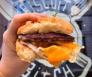 A hand holds a biscuit sandwich filled with bacon, egg, and melted cheese in front of a window with a restaurant logo.