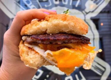 A hand holds a biscuit sandwich filled with bacon, egg, and melted cheese in front of a window with a restaurant logo.