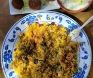 A plate of spiced rice with meat, a side of falafel on lettuce with dip, and a bowl of yogurt topped with spices on a table.