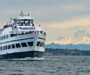 A white Argosy Cruises ferry with passengers sails near Seattle, with a snow-capped mountain and cityscape visible in the background.
