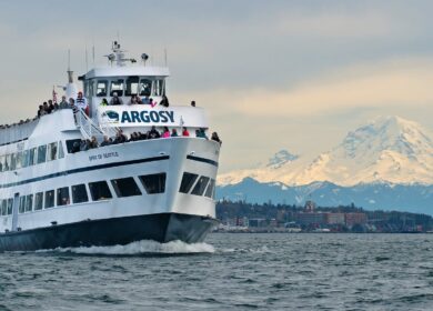 A white Argosy Cruises ferry with passengers sails near Seattle, with a snow-capped mountain and cityscape visible in the background.