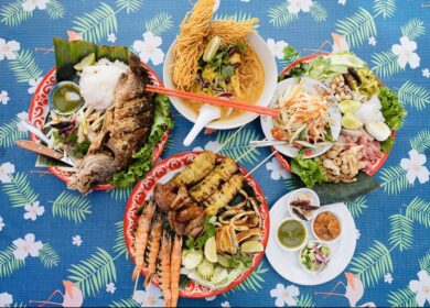 A table set with grilled fish, grilled skewers and prawns, papaya salad, noodle soup, sauces, and fresh vegetables on a blue floral-patterned tablecloth.