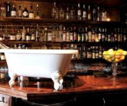 A white clawfoot bathtub sits on a polished wooden bar counter, with shelves of liquor bottles and a bowl of lemons in the background.
