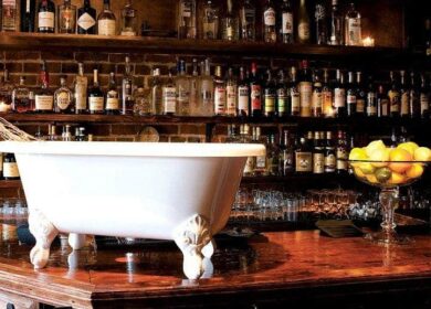 A white clawfoot bathtub sits on a polished wooden bar counter, with shelves of liquor bottles and a bowl of lemons in the background.