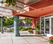 Entrance to Belltown Inn with glass doors, flower pots, decorative columns, and awnings, located on a sunny sidewalk with greenery in the background.