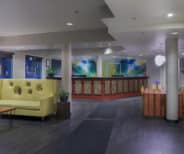 Modern hotel lobby with a yellow sofa, coffee table, reception desk, indoor plants, artwork on the back wall, and dark wood flooring.
