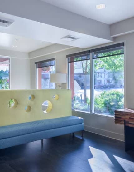 A modern waiting area with a blue bench, a yellow divider with circular cutouts, large windows, and a wooden table with a potted plant.