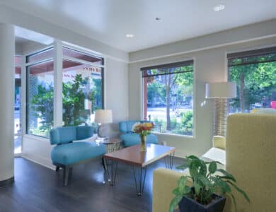 A modern lobby features blue sofas, a yellow chair, a coffee table with flowers, large windows with views of greenery, and indoor plants.
