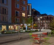 Outdoor seating area with colorful chairs and tables on a city sidewalk at dusk, with buildings and a neon “Belltown Inn” sign in the background.