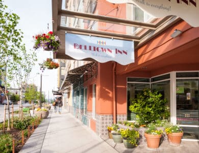 The exterior of Belltown Inn features a covered entrance, potted plants, hanging flower baskets, and a sidewalk on a sunny day.