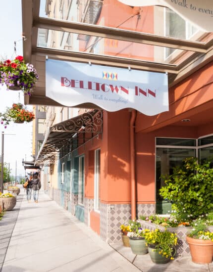 The exterior of Belltown Inn features a covered entrance, potted plants, hanging flower baskets, and a sidewalk on a sunny day.