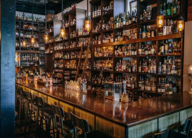 A bar with a long wooden counter, high chairs, and shelves filled with bottles of liquor under warm hanging lights.