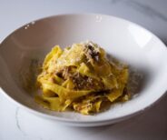 A white bowl filled with tagliatelle pasta in a meat sauce, topped with grated cheese, on a white marble surface.