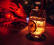 A glass of sparkling beverage with a lemon slice on a coaster sits beside a plate of food in a dimly lit, red-toned setting.