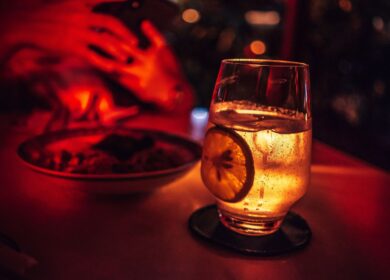 A glass of sparkling beverage with a lemon slice on a coaster sits beside a plate of food in a dimly lit, red-toned setting.