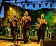 A band performs on stage at night with multiple musicians playing saxophones and guitar under green and yellow stage lights.