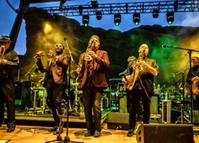 A band performs on stage at night with multiple musicians playing saxophones and guitar under green and yellow stage lights.