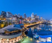 A waterfront view of downtown Seattle at dusk, featuring city buildings, a marina with docked boats, and the illuminated Seattle Great Wheel.