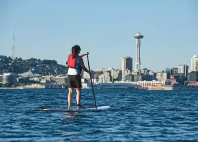 A person wearing a red life vest paddleboards on a body of water with the Seattle skyline and Space Needle visible in the background.