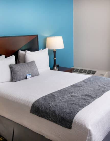 A neatly made bed with white linens, gray accents, and four pillows in a modern hotel room, next to a nightstand with a lamp and a window with yellow curtains.