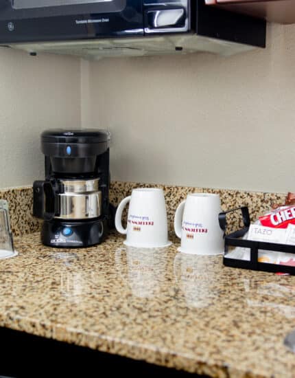 A hotel room countertop with a coffee maker, two upside-down glasses, two coffee mugs, and a tray holding tea bags and Cheez-It snacks next to a sink.