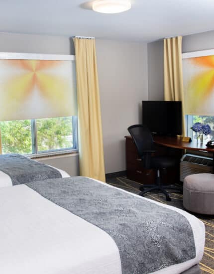Hotel room with two double beds, a desk with a computer monitor, a chair, an ottoman, and two windows with yellow curtains and patterned shades.
