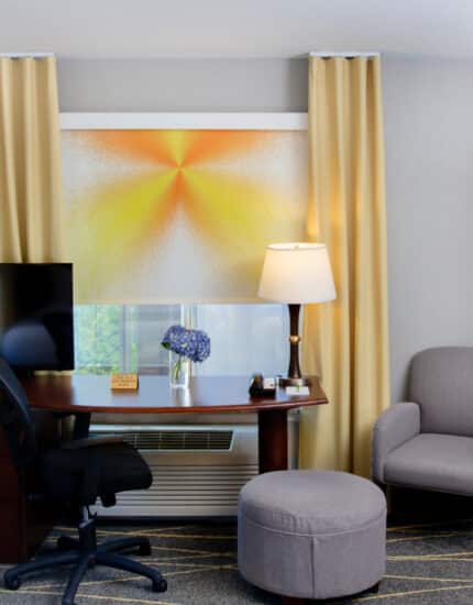 A hotel room workspace with a desk, office chair, TV, lamp, flowers, armchair, and ottoman, with yellow curtains and abstract window shade.