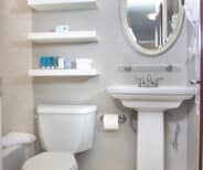 A bathroom with a toilet, pedestal sink, oval mirror, wall-mounted shelves with towels and toiletries, and a wall-mounted hair dryer.