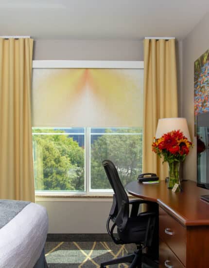 A hotel room with a bed, desk, office chair, TV, lamp, vase of flowers, and a window with yellow curtains and a view of trees outside.