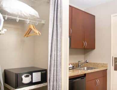 A hotel room kitchenette with a sink, mini fridge, upper cabinets, and a closet area containing a safe, hangers, ironing board, and pillow.