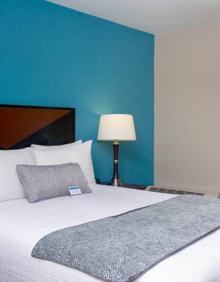 A neatly made bed with white linens and gray accents is next to a gray armchair, a floor lamp, and a window with yellow curtains in a modern hotel room.