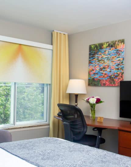 Modern hotel room with a bed, desk, office chair, flat-screen TV, lamp, armchair, window with yellow curtains, and wall art above the desk.