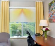 A modern office space with a window, yellow curtains, a desk with a lamp, flowers, phone, and chair, and a gray armchair next to a colorful painting on the wall.