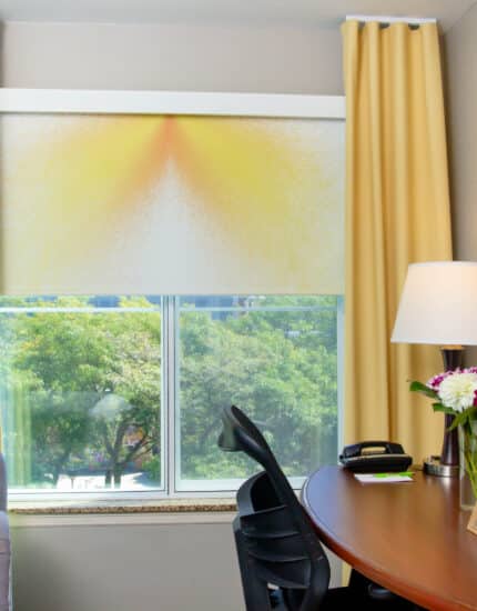 A hotel room with a gray armchair, a desk with a lamp, flowers, and a phone, a window with yellow curtains, and a colorful painting on the wall.