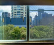 View through a window showing green trees in the foreground and several modern high-rise buildings under a partly cloudy sky in the background.