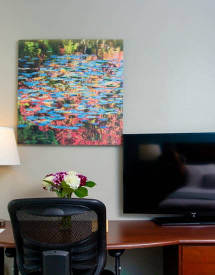 A desk with a black chair, a lamp, a vase of flowers, a flat-screen TV, and a colorful abstract painting on the wall above.