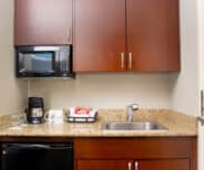 A small kitchenette with granite countertop, sink, coffee maker, mugs, snacks, microwave, and mini fridge beneath brown cabinets.