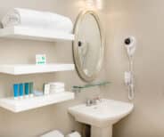 A bathroom with a mirror, sink, wall-mounted shelves holding towels and toiletries, a hairdryer, and a towel bar on a beige wall.