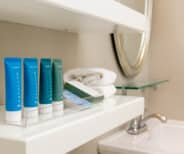 Four hotel toiletry bottles and a box are arranged on a white bathroom shelf next to folded towels, with a sink and mirror visible in the background.