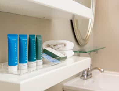 Small bottles of hotel toiletries and a box are arranged on a bathroom shelf next to folded white towels, with a sink and mirror visible nearby.