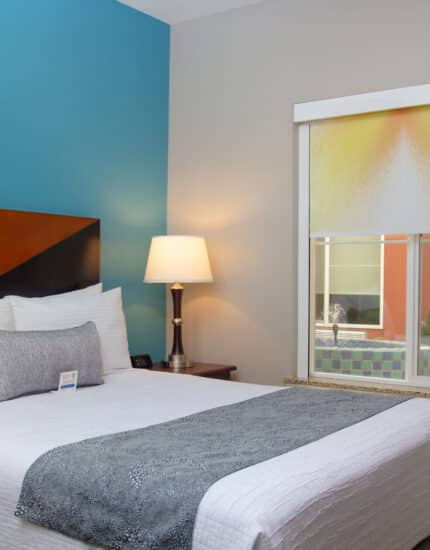 A neatly made bed with white linens and gray accents in a hotel room with a teal accent wall, a lamp, window, and closed door.