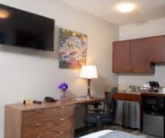 Hotel room corner with a wall-mounted TV, a wooden desk with a lamp and chair, a dresser, a kitchenette with microwave, mini fridge, coffee maker, and a colorful painting above the desk.