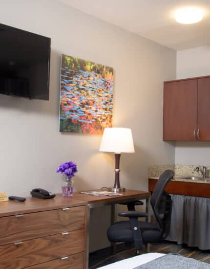 Hotel room workspace with a desk, office chair, lamp, dresser, flat-screen TV, wall art, mini fridge, coffee maker, and upper cabinets.