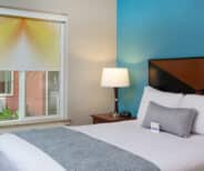 A neatly made bed with white linens and a gray pillow sits next to a bedside table with a lamp. A window with a yellow patterned shade is on the left wall.