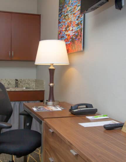 A hotel room workspace with a wooden desk, office chair, lamp, phone, tissues, TV remote, and a kitchenette area with a coffee maker and mini fridge in the background.