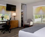 Modern hotel room with a bed, desk and chair, dresser with lamp, armchair, mounted TV, and two large windows with roller shades.