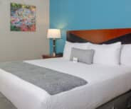 A neatly made bed with white linens and a gray throw blanket, flanked by bedside tables, a lamp, and a colorful wall painting above.