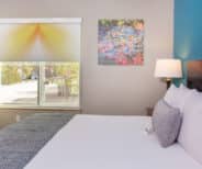 A neatly made bed with white linens, a gray pillow, and a dark headboard is beside a lit lamp, a window with a patterned shade, an armchair, and colorful wall art.